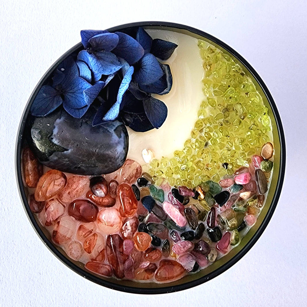 The 8oz Soy Crystal Intention Candle features indigo gabbro, fire quartz, mixed tourmaline, and peridot with a cassia and vanilla scent; it is hand-poured with 100% American soy wax and phthlate-free essential oils and fragrance